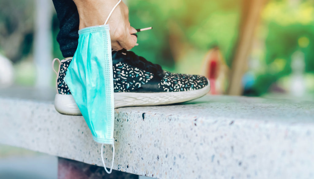 Fitness woman take off her surgical mask getting ready to do run exercise lacing running shoes for outdoor jogging. New normal lifestyle. Health care concept. Selective focus on surgical mask.